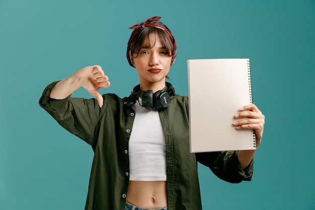 Unpleased young student girl wearing bandana sunglasses and headphones around neck showing large note pad looking at camera showing thumb down isolated on blue background