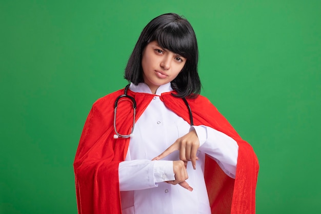 Free photo unpleased young superhero girl wearing stethoscope with medical robe and cloak showing wrist clock gesture isolated on green wall