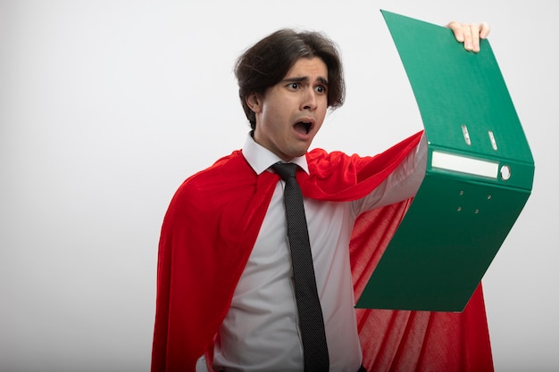 Unpleased young superhero guy wearing tie holding and looking at clipboard isolated on white background