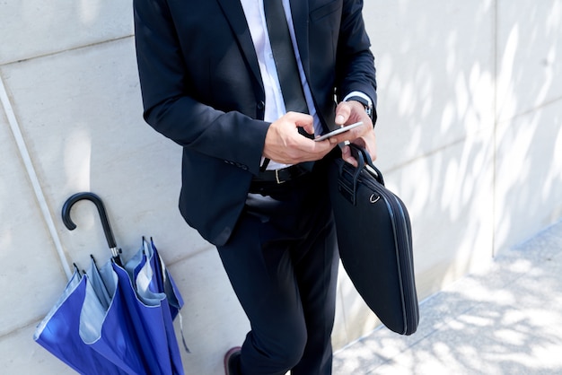 Unrecognizable businessman with briefcase and umbrella standing outdoors and using smartphone