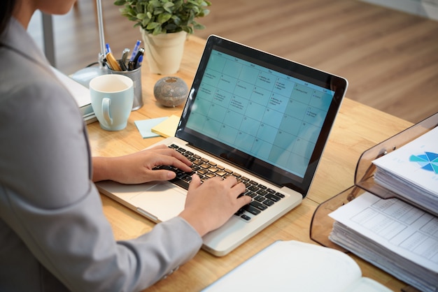 Free Photo unrecognizable businesswoman sitting at desk with laptop and looking at calendar