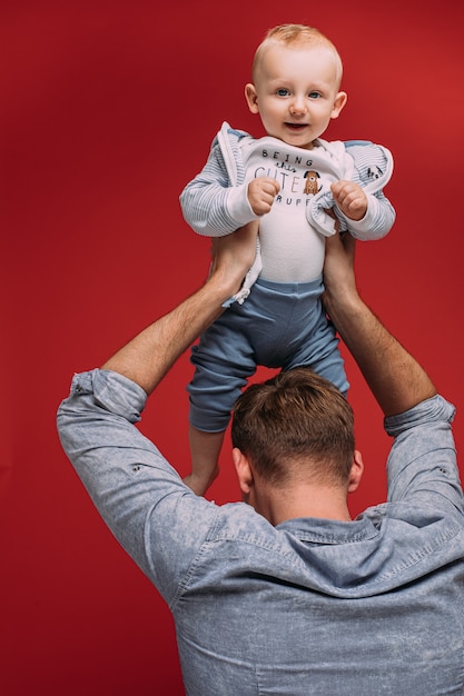 Free photo unrecognizable dad holding his cute baby son up in arms above his head against red background. smiling baby boy looking at camera.