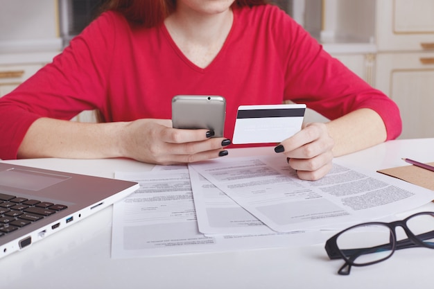 Unrecognizable female model in red sweater sits at working table surrounded with papers and laptop computer