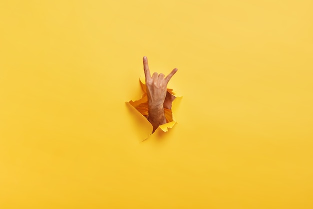 Free Photo unrecognizable man makes rock n roll gesture through ripped hole in yellow paper. male demonstrates horn sign with hand stretched in gap slot of paper. body language concept. colored space