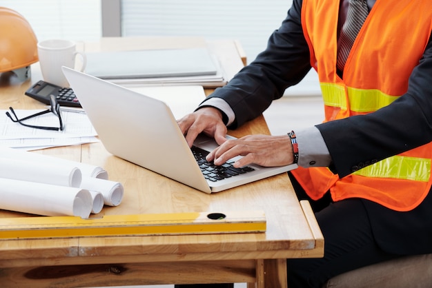 Free photo unrecognizable man in neon safety vest and business suit sitting at desk and using laptop