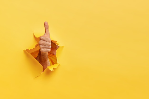 Free Photo unrecognizable man shows like gesture through torn yellow wall, keeps thumb up