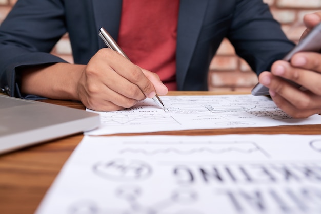 Unrecognizable man sitting at table with smartphone and drawing business diagram on paper