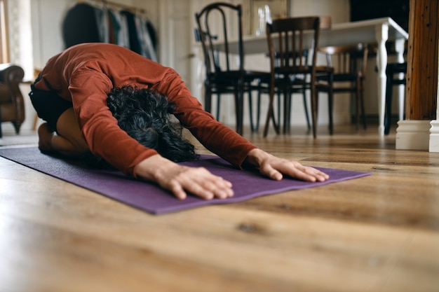 Free photo unrecognizable man with black hair doing yoga at home, having rest in balasana or child pose, relaxing body muscles between asanas, stretching lower back and hips. relaxation and health concept