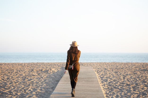 Free photo unrecognizable stylish woman wearing hat, long coat and shoulder bag walking along boardwalk