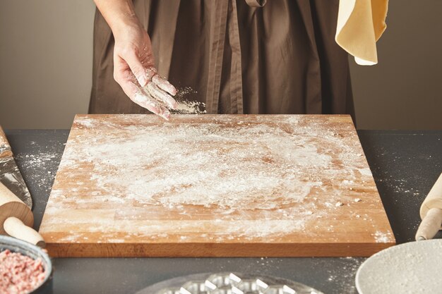 Unrecognizable woman adds some white flour on wooden board while holds flatten dough for pasta or dumplings in air. Ravioli cooking step by step guide