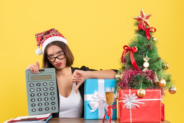 Free photo unsatisfied charming lady in suit with santa claus hat showing calculator making negative gesture in the office on yellow isolated