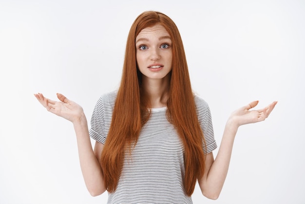 Unsure clueless lovely ginger girl blue eyes wearing summer tshirt shrugging have no idea cannot answer look perplexed questioned raise hands sideways standing white background Copy space