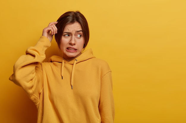 Free photo unsure doubtful woman scratches head, has amnesia, makes tough decision, looks troubled aside, stands puzzled, clenches teeth, dressed in hoodie, poses against yellow wall, empty space for text