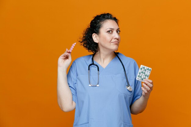 Unsure middleaged female doctor wearing uniform and stethoscope around neck showing packs of pills looking at camera isolated on orange background