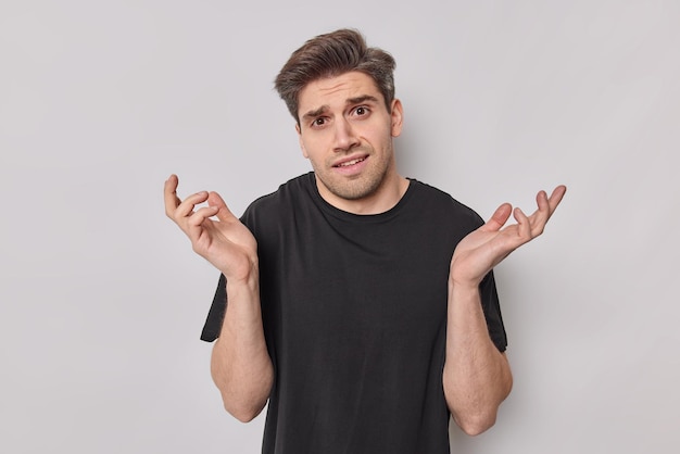 Unsure perplexed young man spreads palms feels doubtful bothered by making choice looks clueless wears casual black t shirt isolated over white background. Questioned male model stands indoor