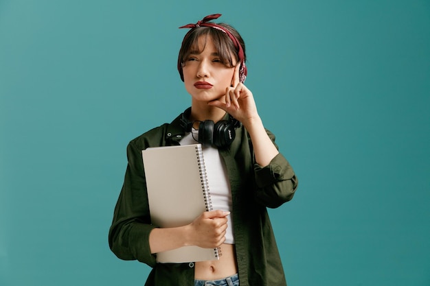 Free photo unsure young student girl wearing bandana and headphones around neck holding large note pad keeping hand on face looking at side thinking isolated on blue background