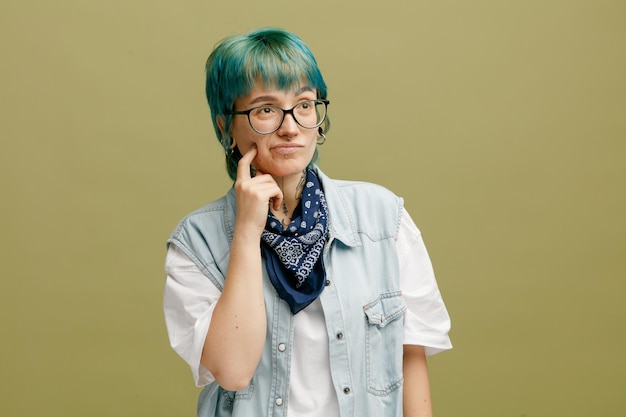 Unsure young woman wearing glasses bandana on neck touching cheek with finger looking at side isolated on olive green background