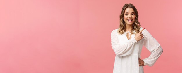 Upbeat goodlooking caucasian blond girl in white dress pointing upper right corner and smiling at ca