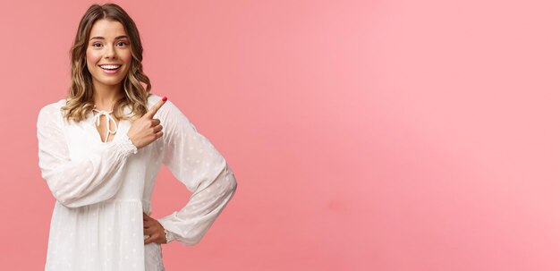 Upbeat goodlooking caucasian blond girl in white dress pointing upper right corner and smiling at camera with interested curious face discuss special promo spring discounts pink background