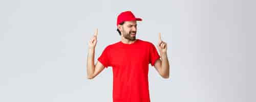 Free photo upbeat handsome bearded delivery guy spending break relaxing employee in red uniform cap and tshirt singing song pointing fingers up carefree close eyes standing grey background