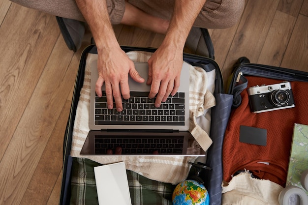 Free photo upper angle view of man sitting on floor with opened suitcase packing for a holiday typing on laptop