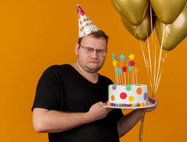 Upset adult slavic man in optical glasses wearing birthday cap holds helium balloons and birthday cake 