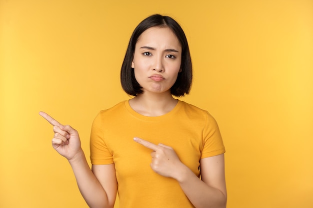 Upset asian woman pointing fingers left showing smth disappointing complaining at banner standing over yellow background