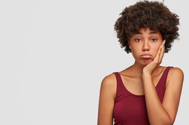 Upset beautiful African American female keeps hand on cheeks, looks displeased