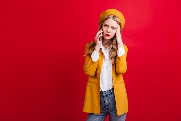 Free photo upset blonde woman in yellow jacket talking on phone. sad caucasian lady in beret standing on red wall with smartphone.