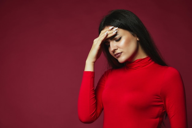 Upset brunette caucasian girl dressed in red pullover with annoying headache put hand on the forehead