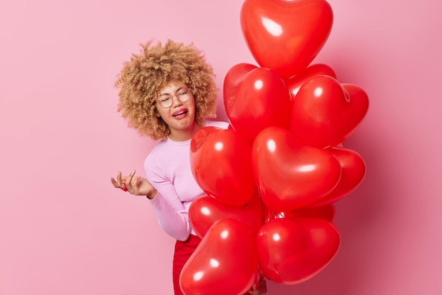 Upset crying woman has spoiled makeup curly hair feels dejected holds bunch of heart balloons keeps hand raised expresses negative emotions isolated over pink background Festive event concept