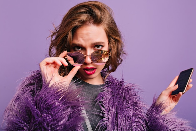 Free photo upset curly woman with smartphone in hand looking through sunglasses on purple background. charming girl in fashionable winter attire with bright make-up posing on photoshoot.