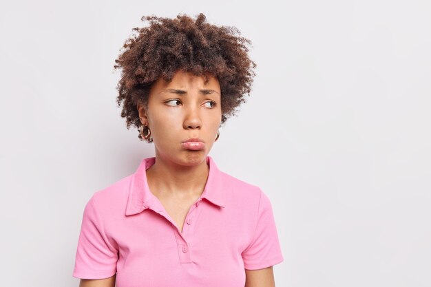 Upset dejected curly haired Afro American woman purses lips looks sadly away has miserable expression dressed in casual pink t shirt poses against white wall