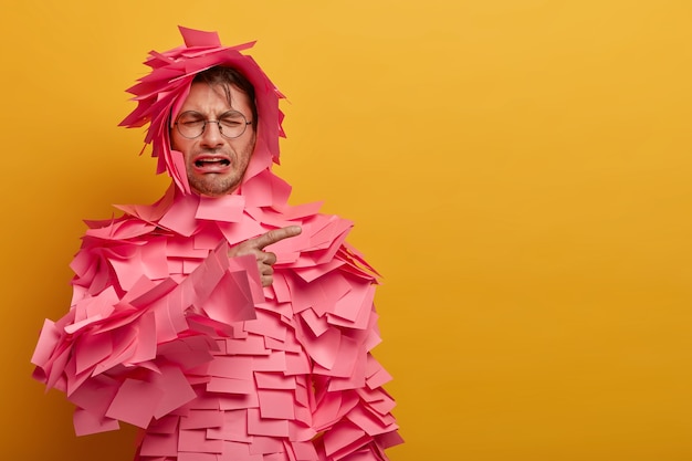 Free photo upset dejected man cries desperately, points away on blank space, dissatisfied with sales discounts, many pink stickers stuck over body, isolated on yellow wall. unhappy desperate feelings