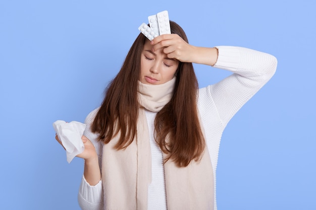 Upset depressed sick woman in warm scarf and casual jumper, holding napkin and pills, frowning with displeased miserable expression, isolated on blue wall, keeps eyes closed.