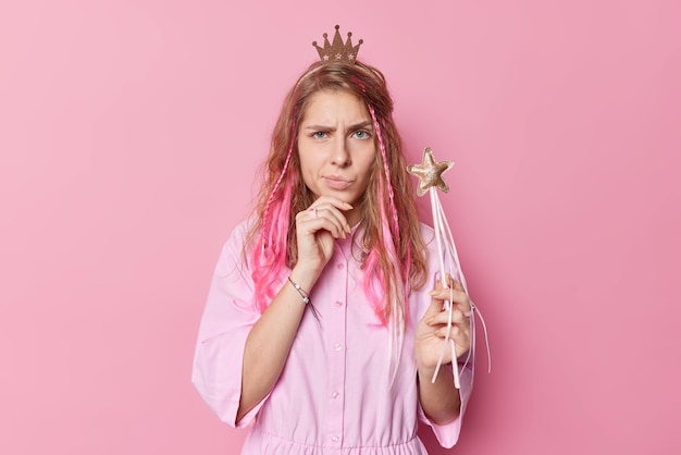 Upset discontent beautiful young woman has sulking expression raises eyebrows wears princess crown and dress holds magic wand ready to make your dreams come true isolated over pink background