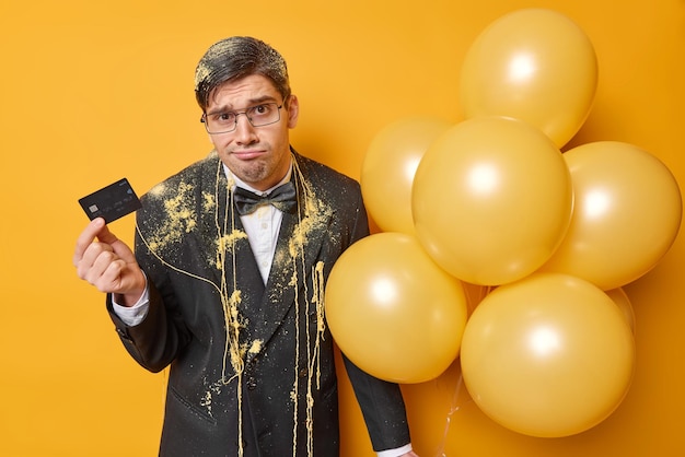 Upset discontent man holds bank ccard has no money for celebration looks sadly purses lips wears formal clothes hods bunch of inflated balloons isolated over yellow background Holidays concept