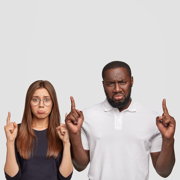 Upset discontent woman and man purse lips in dissatisfaction, point with both index fingers upwards