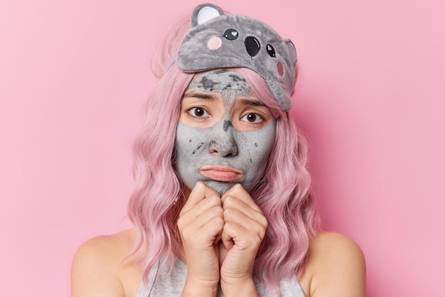 Free photo upset discontent woman with dyed hair keeps hands under chin feels sad applies beauty mask for skin refreshment undergoes face care procedures before sleeping time isolated over pink background