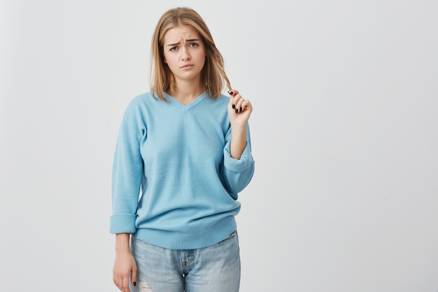 Upset and dissatisfied young woman with oval face, dark eyes and fair straight hair wearing blue casual sweater, frowning her eyebrows and playing with her hair, displeased with something.