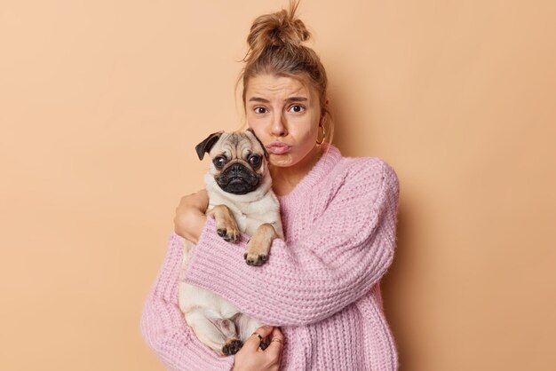 Free photo upset european woman looks sadly at camera embraces pug dog with love feels unhappy because of problems purses lips wears knitted sweater isolated over brown background people and pets concept