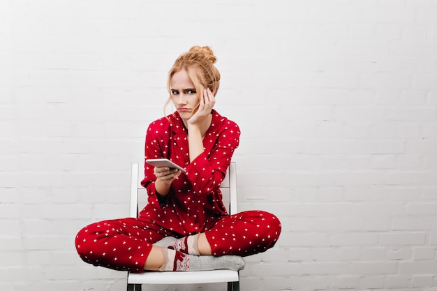 Free Photo upset girl in cute socks sitting on chair and waiting for call. indoor portrait of blonde sad young woman posing with legs crossed and holding smartphone.