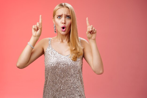 Upset gloomy complaining blond whining girl in silver stylish luxurious dress frowning cringing unhappy pointing up regret jealous tell boyfriend sale over, standing depressed sad red background.