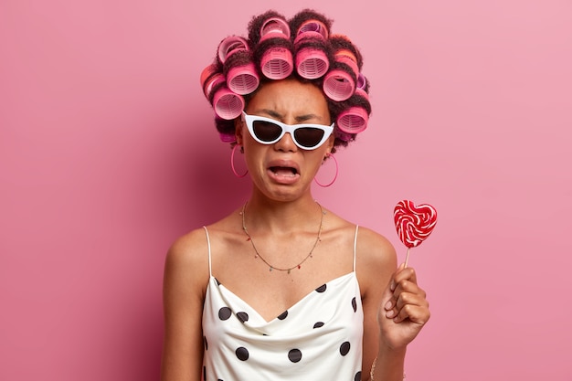 Free photo upset gloomy woman wears hair curlers, cries with displeased expression, gets ready for holiday, makes hairstyle, poses with tasty candy on stick, wears sunglasses and casual dress