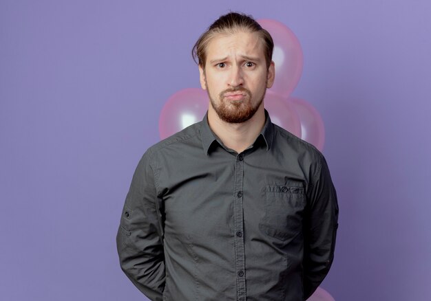 Upset handsome man holds helium balloons behind and looks isolated on purple wall