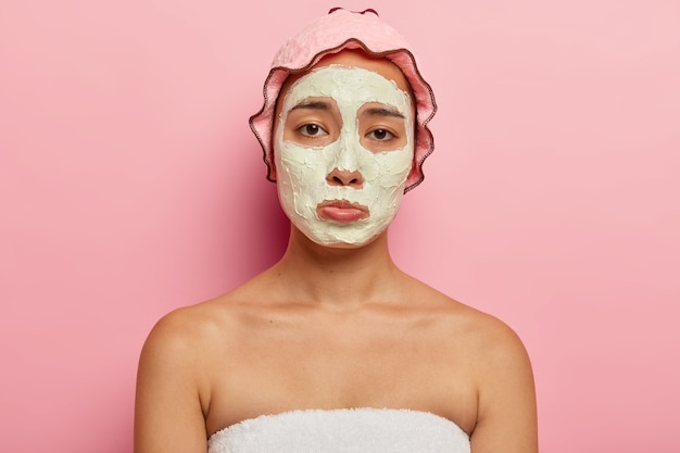 Free photo upset korean young female has dissatisfied sad look, gets beauty treatment, unhappy to have wrinkles and problematic skin, has cosmetic mask on face, wears waterproof headgear for taking shower