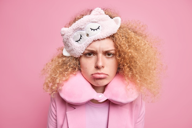 Free photo upset offended beautiful woman with curly hair looks sadly hates early awakening wears sleepmask travel pillow around neck has miserable face expression isolated over pink wall