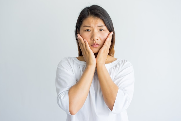 Upset shocked young Asian woman touching cheeks and looking at camera.