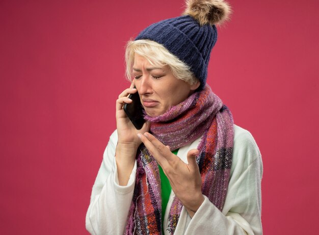 Upset sick unhealthy woman with short hair in warm scarf and hat feeling unwell crying while talking on mobile phone standing over pink wall