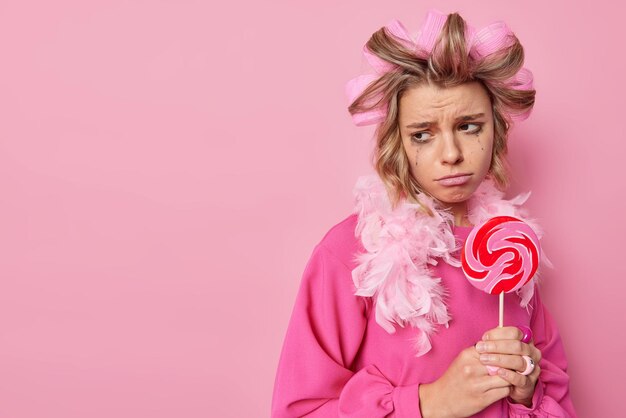 Upset thoughtful woman has leaked makeup holds round lollipop on stick keeps to diet cannot eat sweets applies hair rollers for making haircut wears festive dress poses against pink background